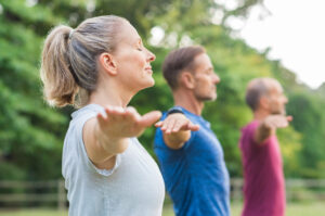 Group of senior people with closed eyes stretching arms at park. Happy mature people doing yoga exercise outdoor on a bright morning. Yoga class with woman and men doing breath exercising with stretched arms. Cenikor’s Aftercare and R